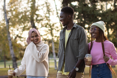 Three friends walk through a park while talking, smiling, and carrying coffee.